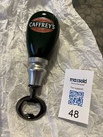 Porcelain Caffreys tap handle with black, red, and white logo. Modified with silver metal neck and black bottle opener at base. Placed on silver crinkled paper.