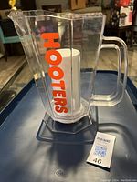 Clear beverage pitcher with handle, orange Hooters logo, and central white ice compartment visible. Shown from side angle on a tray indoors.