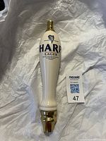 Large white porcelain Harp Lager beer tap handle with blue and black lettering, harp logo, and gold metal accents. Image shows front view of the tap handle on a white background.