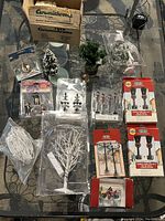View of assorted Christmas village lights, miniature trees, figures, and other accessories spread on a surface, some in packaging.