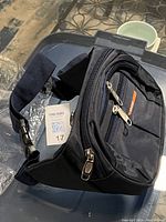 Photo of a new dark blue fanny pack showing its multiple compartments, zippers, and adjustable strap with a plastic buckle, placed on a surface with a MaxSold lot tag nearby.