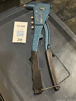 Photo of a handheld rivet tool showing blue metal body with black ergonomic grips, four nozzle tips on the side, and a locking pin at the handle base.