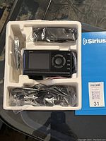 Sirius satellite radio device with screen and circular control pad, remote control, power adapter, and cables in original molded packaging inside blue Sirius-branded box.