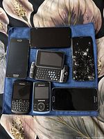 Top view of seven mobile phones on blue cloth; one with cracked screen, mixture of touchscreen and keyboard devices