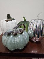 Set of four decorative pumpkins shown on a wooden surface, featuring white ceramic, matte green, silver-toned glass, and speckled teal glass pumpkins.