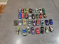 Top view of 32 assorted diecast vehicles including cars, motorcycles, and aircraft models arranged in rows on a grey surface.