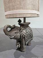 Close-up of the elephant lamp base showing detailed carved traditional patterns and the attached beige textured lampshade with fabric trim and decorative square finial.