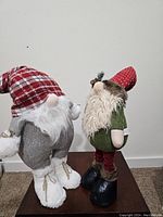 Side view of two holiday gnomes, one wearing red plaid hat with herringbone pants, and the other in green with pinecone-adorned hat