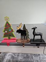 Three colorful metal stocking holders: Christmas tree with button details, moose with forest backdrop, black deer.