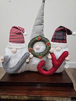 Photo of three festive holiday gnomes holding letters J, O (wreath), Y spelling JOY, arranged side by side on a wooden surface.