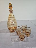 Photo of amber glass decanter with gold striped detailing and four matching shot glasses arranged in front on a white surface, showing the set from a right angle