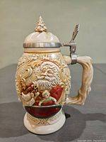 Front and side view of the vintage Avon Father Christmas stein showing detailed sculpted holiday scenes, red and green color accents, tree branch style handle, and pinecone-shaped lid finial.