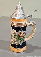 Side view of the beer stein showing detailed ceramic relief artwork depicting alpine cottages, mountains, and floral designs with pewter lid closed.