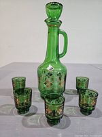 Image of the green glass decanter with handle and stopper surrounded by six matching shot glasses, all decorated with gold accents