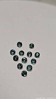 Thirteen round cut sapphires showing aqua blue with teal tones scattered on a white surface.