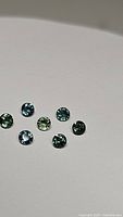 Seven loose round cut sapphires in blue, teal, and bright green arranged on white background, clearly showing facets and color variations.