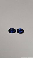 Pair of oval cut royal blue sapphire gemstones shown from above on neutral background.