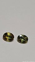 Close-up photo of two oval faceted natural sapphires showing yellow-green-blue tri-color shades on a neutral background.