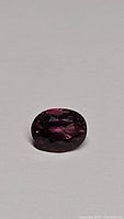 Close-up photo of an oval cut sapphire gemstone exhibiting deep hot pink to purple coloring under lighting conditions, placed on neutral background