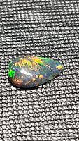Pear-shaped cabochon black opal gemstone resting on textured surface showing vibrant multi-color fire including green, orange, red, and yellow hues.