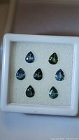 Seven pear-shaped multicolored sapphires with blue, green, and yellow hues in a display case on white foam backing.