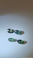 Four oval natural sapphires, green with blue undertones, on white background under lamp light showing faceting and color.