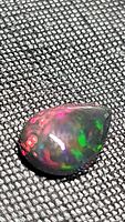 Close up image of pear-shaped black opal showing strong rainbow flash with dominant red and green tones on a dark background.