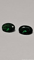 Close-up photo showing two oval vivid green tsavorite garnet gemstones resting on a neutral background under even light, displaying their size, shape, and color.