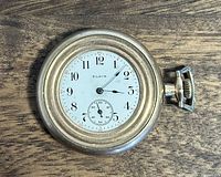 Front face of Elgin gold pocket watch showing white dial with black Arabic numerals and sub-second dial, gold-tone case, winding crown on the side.