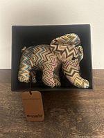 Soft fabric elephant plush in colorful zigzag pattern inside a black box with Holt Renfrew tag and OrphanAid Africa branding.