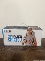 Front view of Homedics Quad Action Shiatsu massager box, showing male model holding massager on shoulders and neck area with straps.