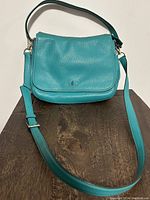Full front of teal leather handbag with strap attached, displaying the flap with the small gold Kate Spade logo at the center.