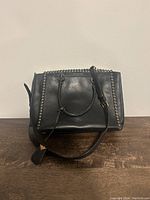 Front view of black Michael Kors leather handbag with studded edges and dual handles lying on a wooden surface.