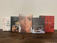 Five hardback coffee table books arranged side by side on a wooden surface. Titles visible: 'Yes Way Rosé,' 'Cooking for Cocktails,' 'Face Forward,' 'Vancouver Eats,' 'Toppino's.'