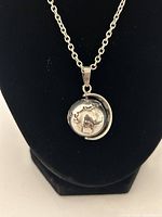 Close-up photo of silver globe atlas pendant hanging on chain against a black background, showing details of continents and globe arm.