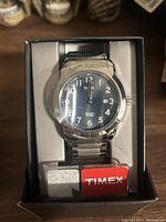 Front view of new Timex wristwatch in original box showing blue dial with white numerals, date window, stainless steel bracelet, and Indiglo tag