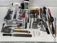 Wide view showing entire lot of kitchen utensils spread on a white cloth background. Items include graters, peelers, spatulas, openers, whisks, scoops, measuring spoons, and more.