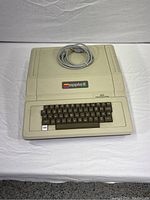 Front photo of vintage Apple II Plus computer showing beige plastic housing, keyboard, and coiled AC cord on top.