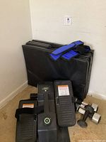 Cubii exercise machine with foot pedals, workout mat folded in corner, dumbbells and blue ankle weights arranged on floor