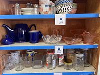 Shelf display showing royal blue teapot with lid, two matching coffee mugs, clear glass bowl, pink glass candy dish and other glassware items