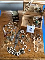 Wide view of costume jewelry lot on wooden table showing pearl necklace draped over jewelry box, brooches, cufflinks, earrings and bracelets.