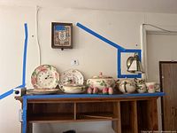 Shelf display showing English china dishware set with pink flowers: plates, teapot, coffee pot, mugs, salt and pepper shakers, and two ceramic clocks, one matching the china design. Also includes wooden shelf backing and blue tape along edges.