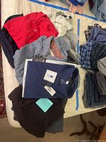 Assorted men's clothing laid on bed including red collared shirt, white lace shirt, grey sweatpants, and navy blue shirt with tags
