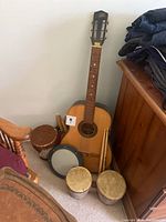 Acoustic guitar resting with bongo drum, two handheld drums, drumsticks and four ping-pong paddles near a wooden cabinet and chair.