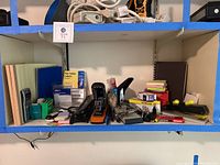Shelf holding various office supplies including legal pads, staplers, tape dispensers, hole punch, notebooks, file folder labels, and other items.