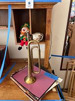 Stuffed leprechaun doll, Peter Rabbit mug and creamer, brass trumpet, and a stack of photos with blue tape measurement tag showing 11.5x14.5 inches approx.