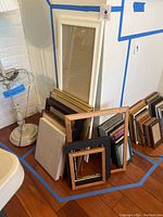 Photo showing group of picture frames leaning against wall, various sizes and materials including wood, metal, and brass.