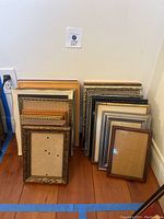 Photo showing collection of 19 picture frames stacked against a wall on a wooden floor. Frames vary in size around 5x7 to 8x10, include wood, metal, gold, brass finishes. Some ornate detailing visible.