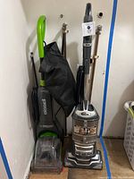 Front view of two upright vacuum cleaners side by side, Bissell on the left with green handle and Shark on the right with metallic gray body, showing overall condition and attachments in black bag.