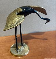 Close up of one brass crane bird statue showing black enamel body, textured brass wings, and round brass base.